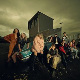 Promotional photo of Atomic! posing on a rooftop car park at dusk, gathered around a scooter and vintage car against a dramatic cloudy sky.