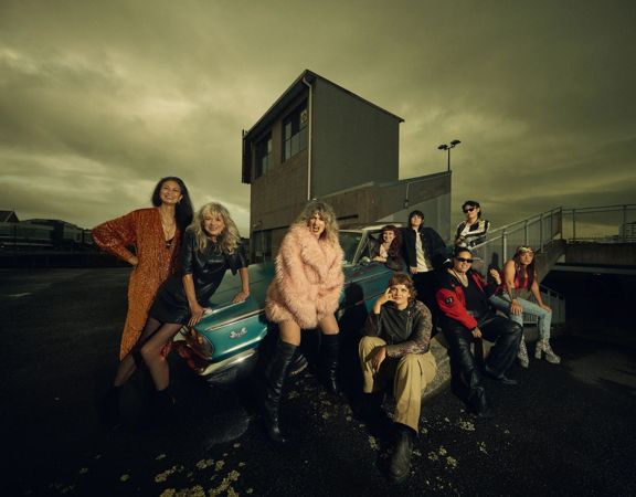 Promotional photo of Atomic! posing on a rooftop car park at dusk, gathered around a scooter and vintage car against a dramatic cloudy sky.