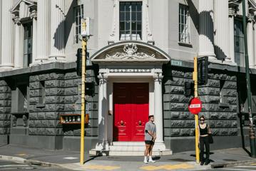 The big red door entrance to Logan Brown, a restaurant on the corner of Cuba Street and Vivian Street in Te Aro, Wellington. Two people stand, waiting to cross the street.