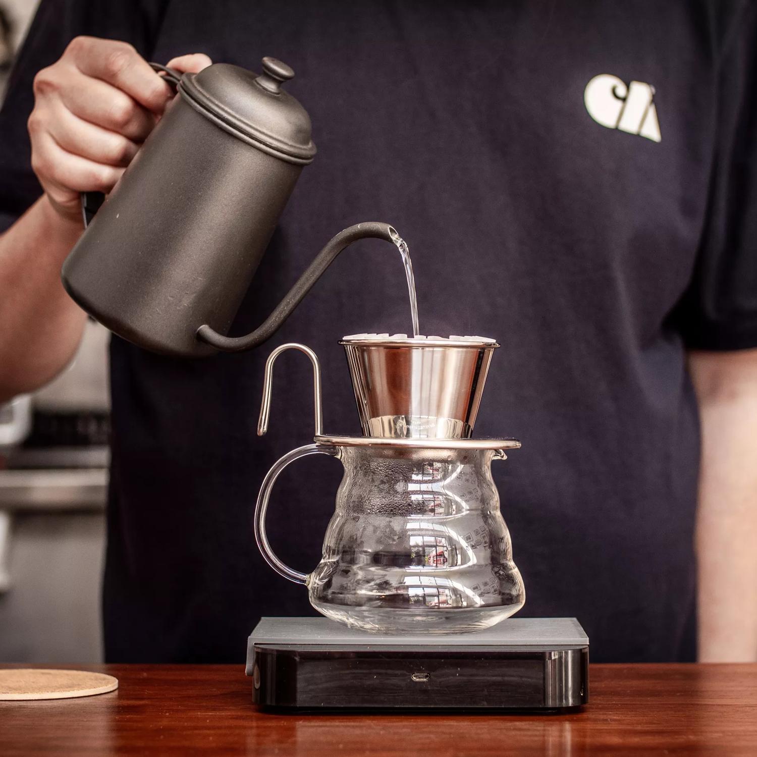 A person pouring boiling water from a black vintage kettle over a coffee filter that's placed on top of a glass jug. 