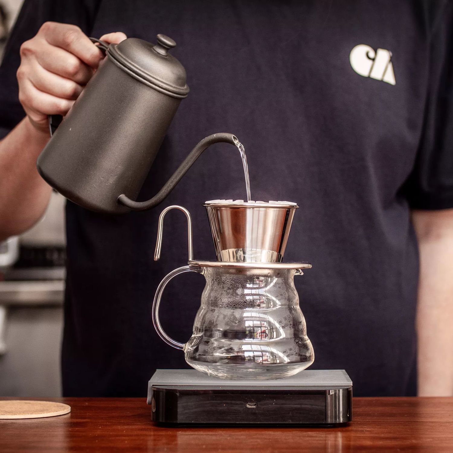 A person pouring boiling water from a black vintage kettle over a coffee filter that's placed on top of a glass jug.