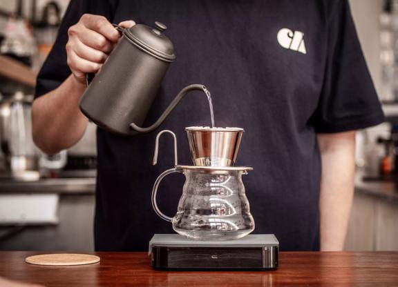 A person pouring boiling water from a black vintage kettle over a coffee filter that's placed on top of a glass jug. 