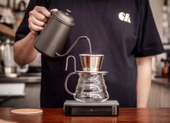 A person pouring boiling water from a black vintage kettle over a coffee filter that's placed on top of a glass jug.