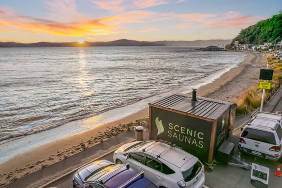 A Scenic Sauna cabin sauna parked on the beach in Days Bay, Eastbourne. The sun is setting over the hill.