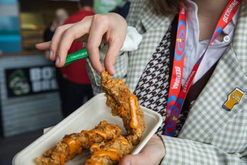 A hand picks up one of three chicken tenders, drizzled with orange sauce, from a takeaway container.