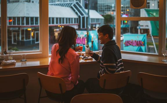 Two people enjoy food and drinks sitting at a table next to the window at Supra.