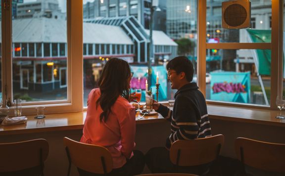 Two people enjoy food and drinks sitting at a table next to the window at Supra.