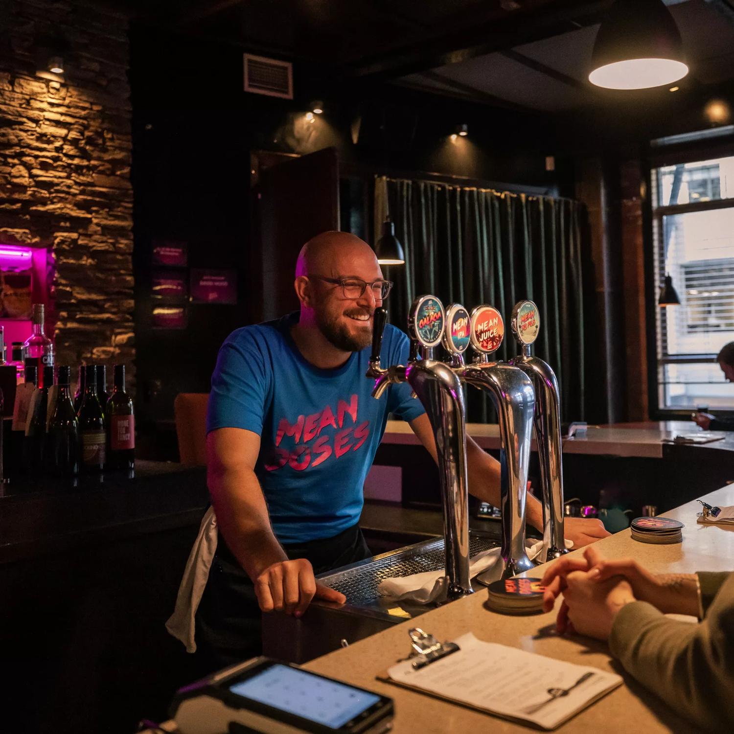 A smiling bartender in a blue teeshirt at Mean Doses stands behind the bar taking an order.