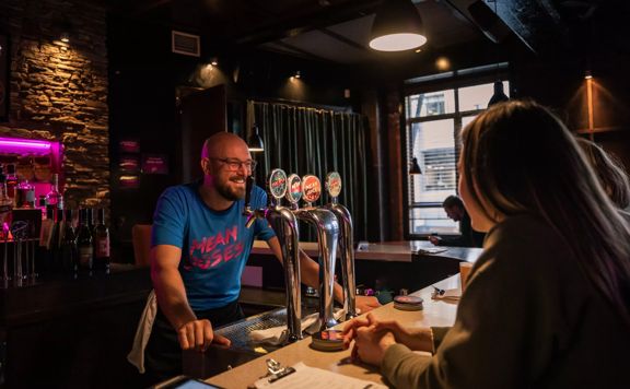 A smiling bartender in a blue teeshirt at Mean Doses stands behind the bar taking an order.