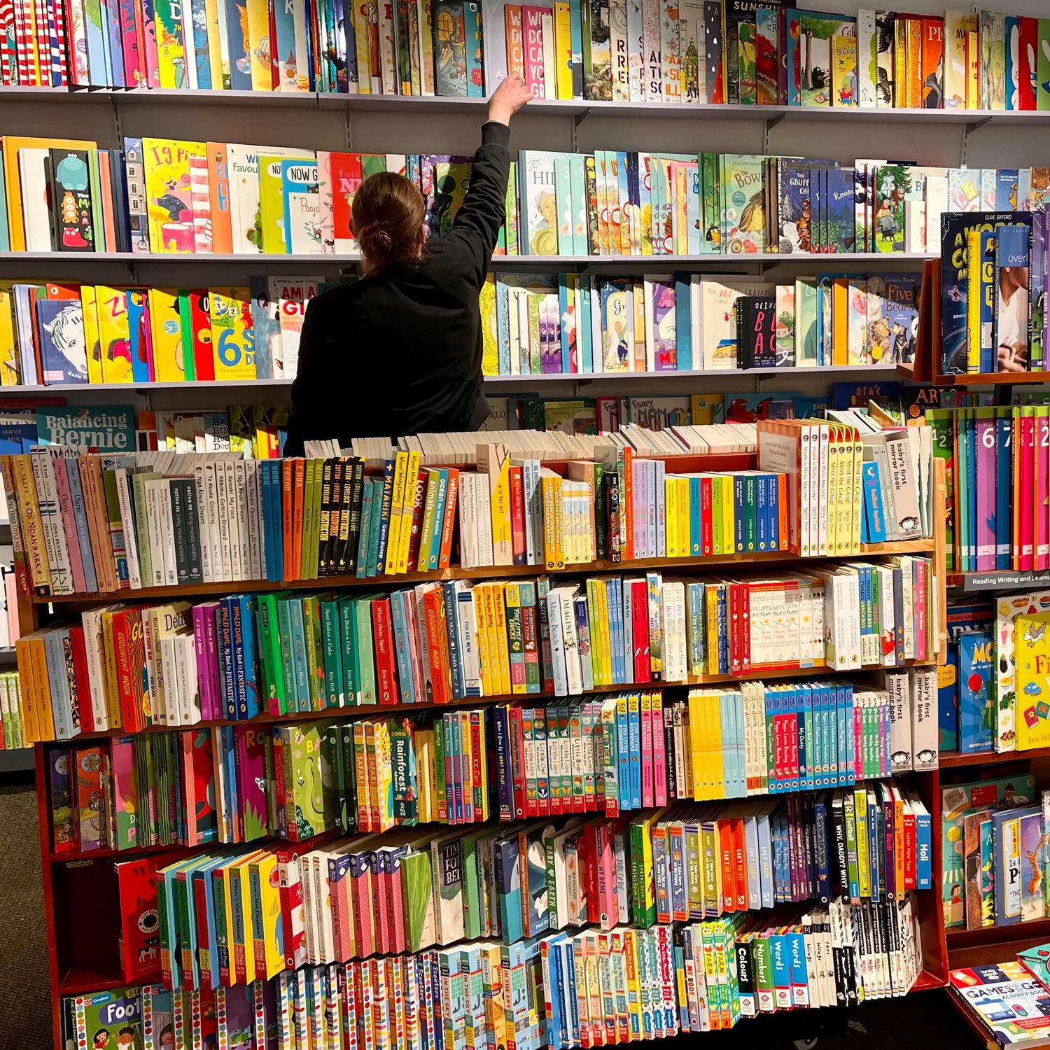 Inside Marsden Books, a small bookstore in Karori Wellington, a person facing away reaches for a book on a high shelf. The shelves in this corner of the store are full of colourful children's literature.