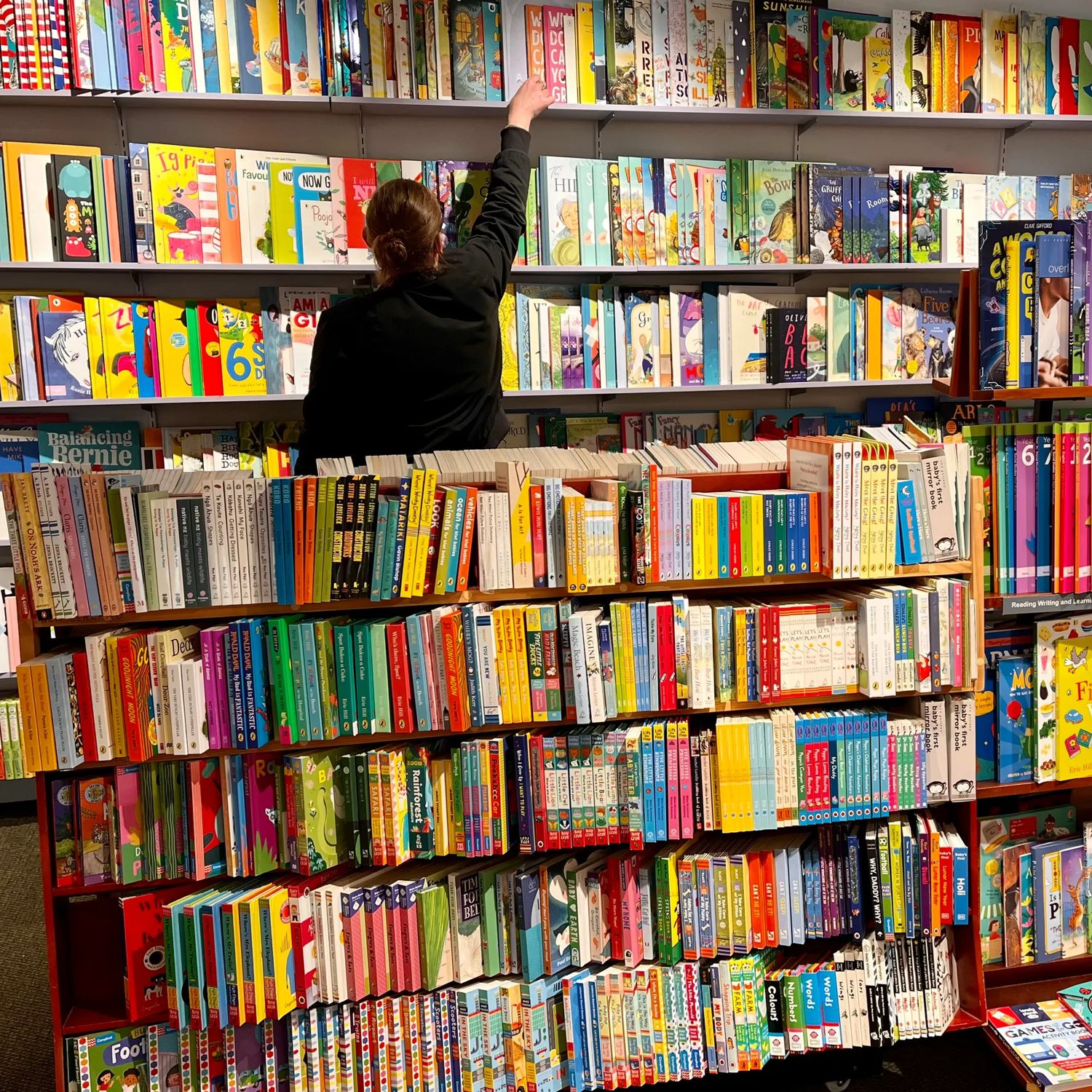 Inside Marsden Books, a small bookstore in Karori Wellington, a person facing away reaches for a book on a high shelf. The shelves in this corner of the store are full of colourful children's literature.