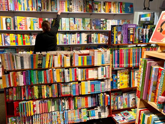 Inside Marsden Books, a small bookstore in Karori Wellington, a person facing away reaches for a book on a high shelf. The shelves in this corner of the store are full of colourful children's literature.