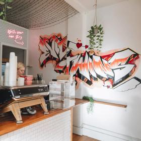 A barista at Evil Twins cafe on Willis Street, making coffee behind the counter. A large graffiti is on the wall that says Evil Twins.