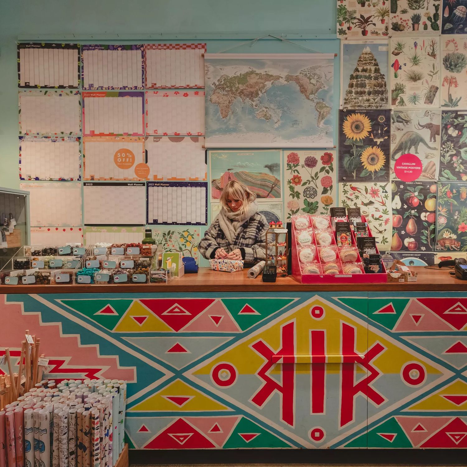The vibrant interior of Iko Iko with a staff member wrapping presents. Celndars and posters adorn the walls and the counter is painted with red, blue and yellow triangles.