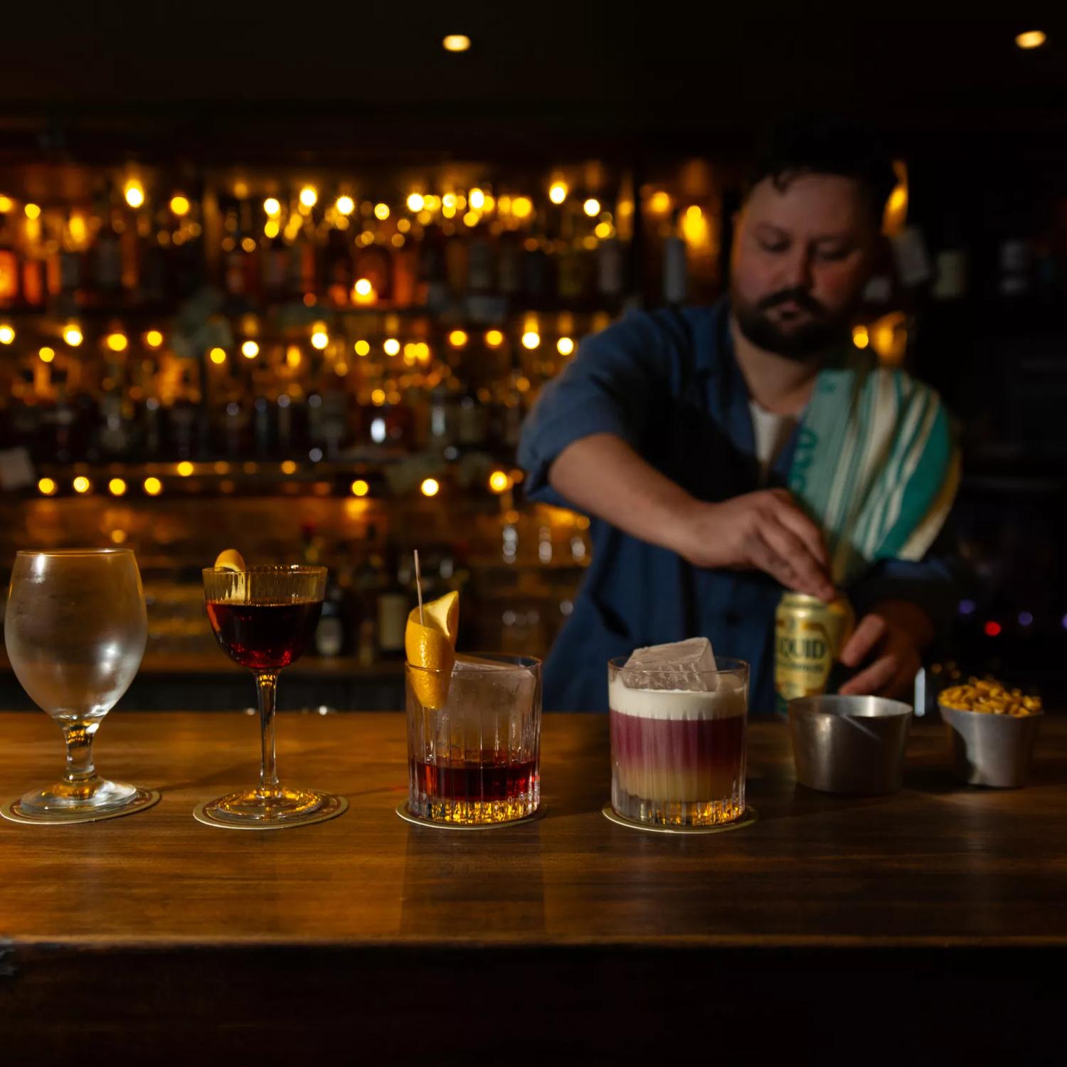 Three cocktails and a beer glass are lined up on a wooden bar at Dee's Place located on Cuba Street at Ghuznee Street. A bartender, with a rag on his shoulder is opening a can of beer behind the bar. 