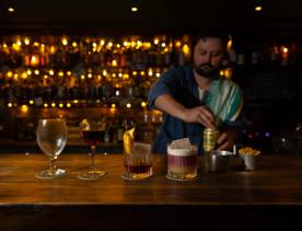 Three cocktails and a beer glass are lined up on a wooden bar at Dee's Place located on Cuba Street at Ghuznee Street. A bartender, with a rag on his shoulder is opening a can of beer behind the bar.