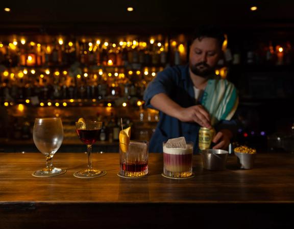 Three cocktails and a beer glass are lined up on a wooden bar at Dee's Place located on Cuba Street at Ghuznee Street. A bartender, with a rag on his shoulder is opening a can of beer behind the bar. 