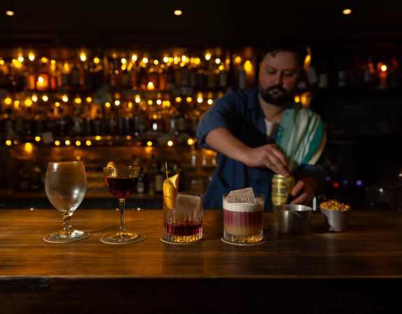 Three cocktails and a beer glass are lined up on a wooden bar at Dee's Place located on Cuba Street at Ghuznee Street. A bartender, with a rag on his shoulder is opening a can of beer behind the bar.