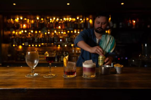 Three cocktails and a beer glass are lined up on a wooden bar at Dee's Place located on Cuba Street at Ghuznee Street. A bartender, with a rag on his shoulder is opening a can of beer behind the bar. 