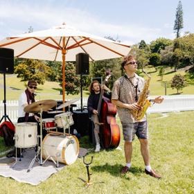 A three-piece band with a drummer, double-bassist and saxophonist play under a sun umbrella in a park.