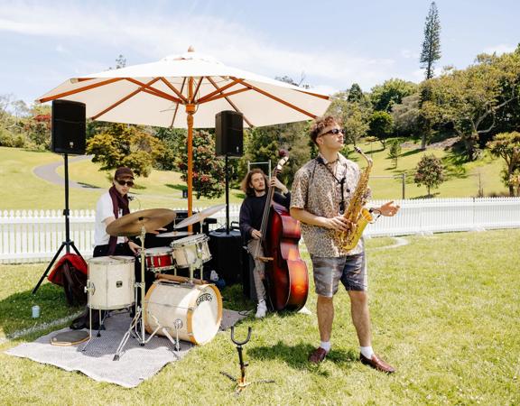 A three-piece band with a drummer, double-bassist and saxophonist play under a sun umbrella in a park.