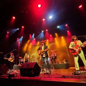A band performs on stage under red, yellow, and blue spotlights with guitars, drums, and speakers visible.