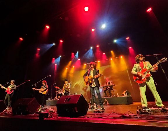 A band performs on stage under red, yellow, and blue spotlights with guitars, drums, and speakers visible.