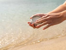 Hands holding an open tin of Seasick Sunscreen on a sandy bead.