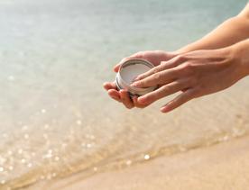 Hands holding an open tin of Seasick Sunscreen on a sandy bead. 