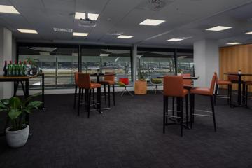 A hotel lounge with tall tables, bar stools, dark grey carpets and three large windows.