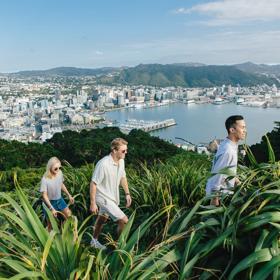 Three people are walking up the trail on Mount Victoria, surrounded by lush greenery, with a view of Wellington city centre behind them. 