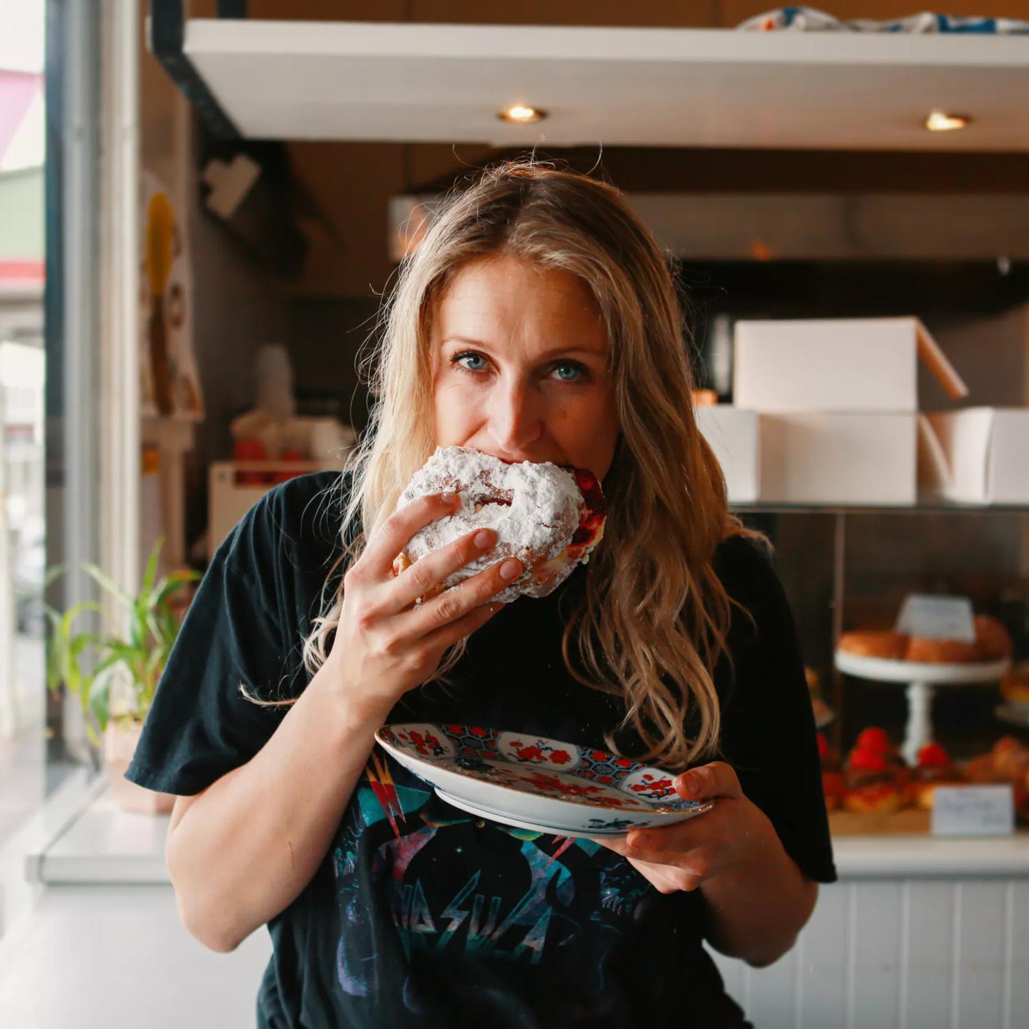 A person wearing a black tee shirt is biting into a powdered pastry and holding a plate in their other hand at Tomboy bakery in  Mount Victoria in Wellington.