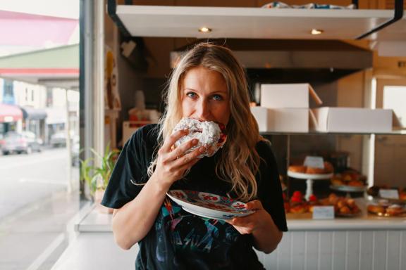 A person wearing a black tee shirt is biting into a powdered pastry and holding a plate in their other hand at Tomboy bakery in  Mount Victoria in Wellington.