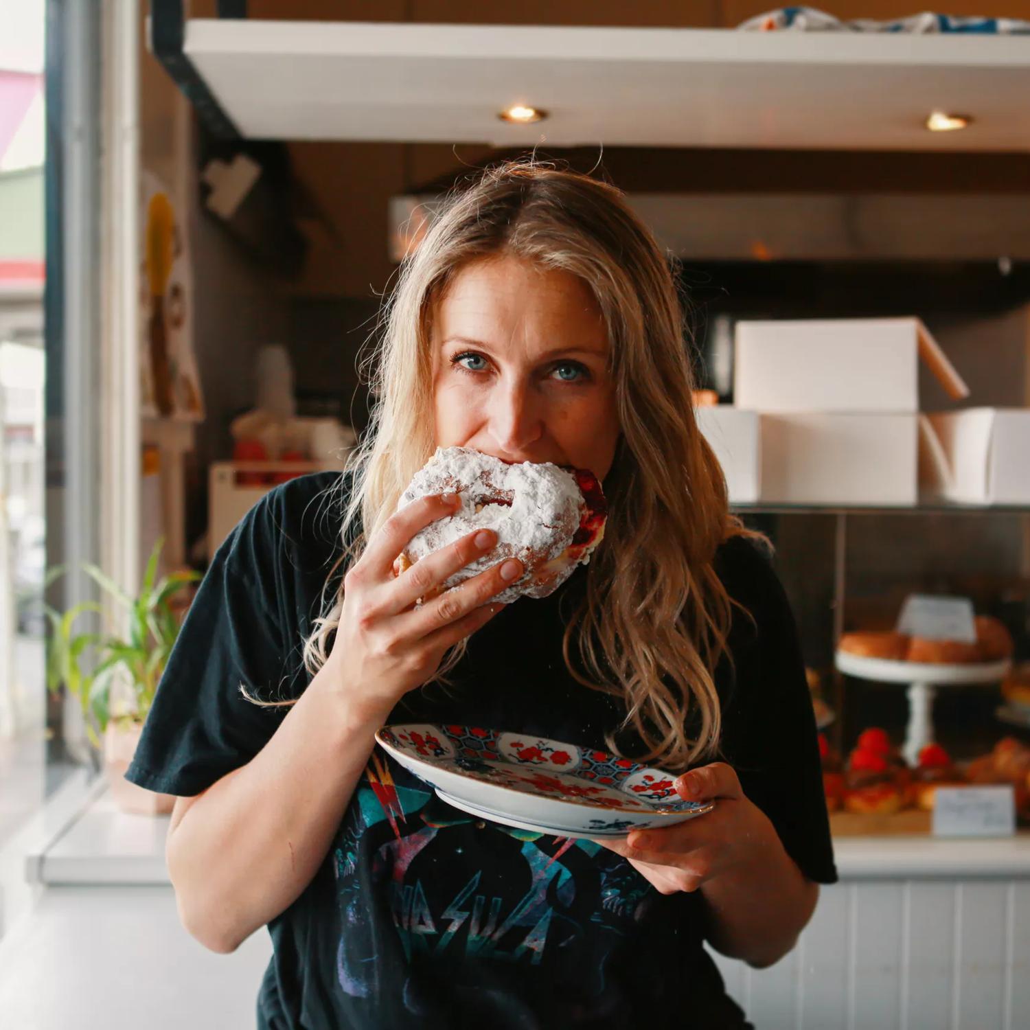 A person wearing a black tee shirt is biting into a powdered pastry and holding a plate in their other hand at Tomboy bakery in  Mount Victoria in Wellington. 