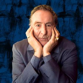 English actor, comedian, songwriter, musician, screenwriter and playwright, Eric Idle wearing a grey suit poses with his face cupped in his hands.