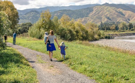 An adult and two children walk along a path in a lush valley with mountains in the background.