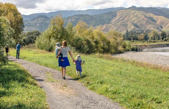An adult and two children walk along a path in a lush valley with mountains in the background.