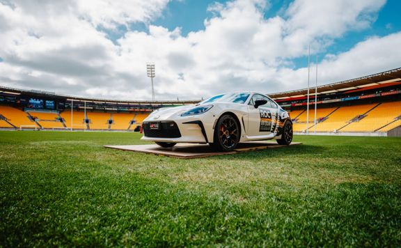 White race car parked on a grass field inside a large stadium with yellow seating and a cloudy sky overhead.