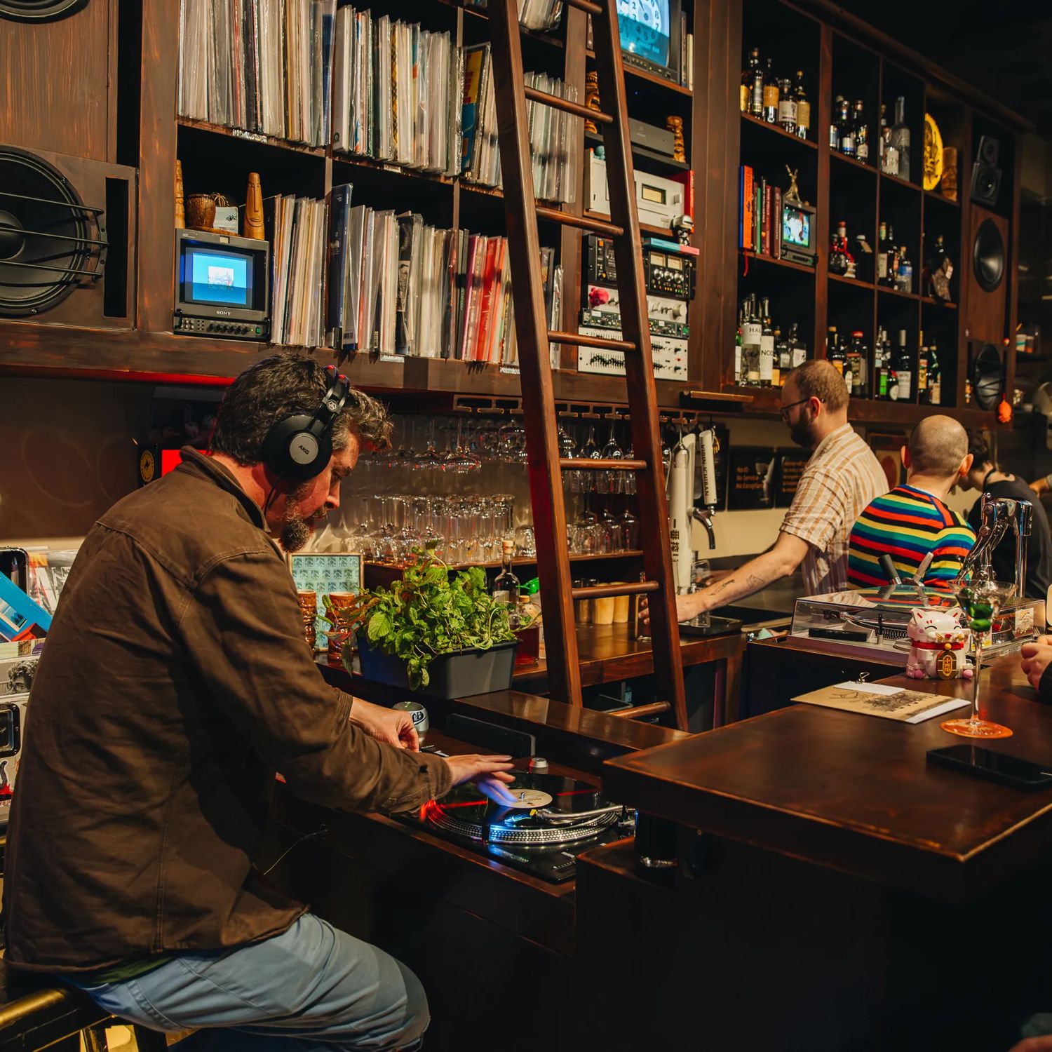 A person wearing headphones operates a turntable at a cozy bar, with shelves of records and bottles behind the counter and several people seated nearby under warm lighting.