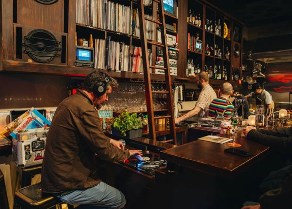 A person wearing headphones operates a turntable at a cozy bar, with shelves of records and bottles behind the counter and several people seated nearby under warm lighting.