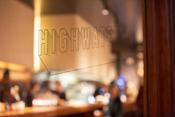 A window at Highwater, a restaurant & wine bar on Cuba Street, Te Aro, Wellington. The restaurant's logo is a decal on the glass and the interior amber lighting shines through the window.