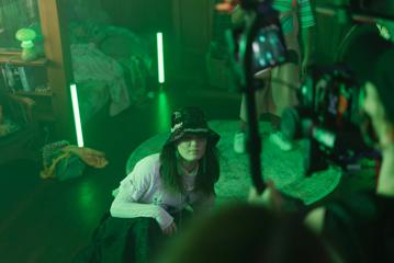 Looking over the shoulder of a camera person filming Little Apocalypse. In front of the camera, a child wearing a bucket hat crouches on the floor in a green room.