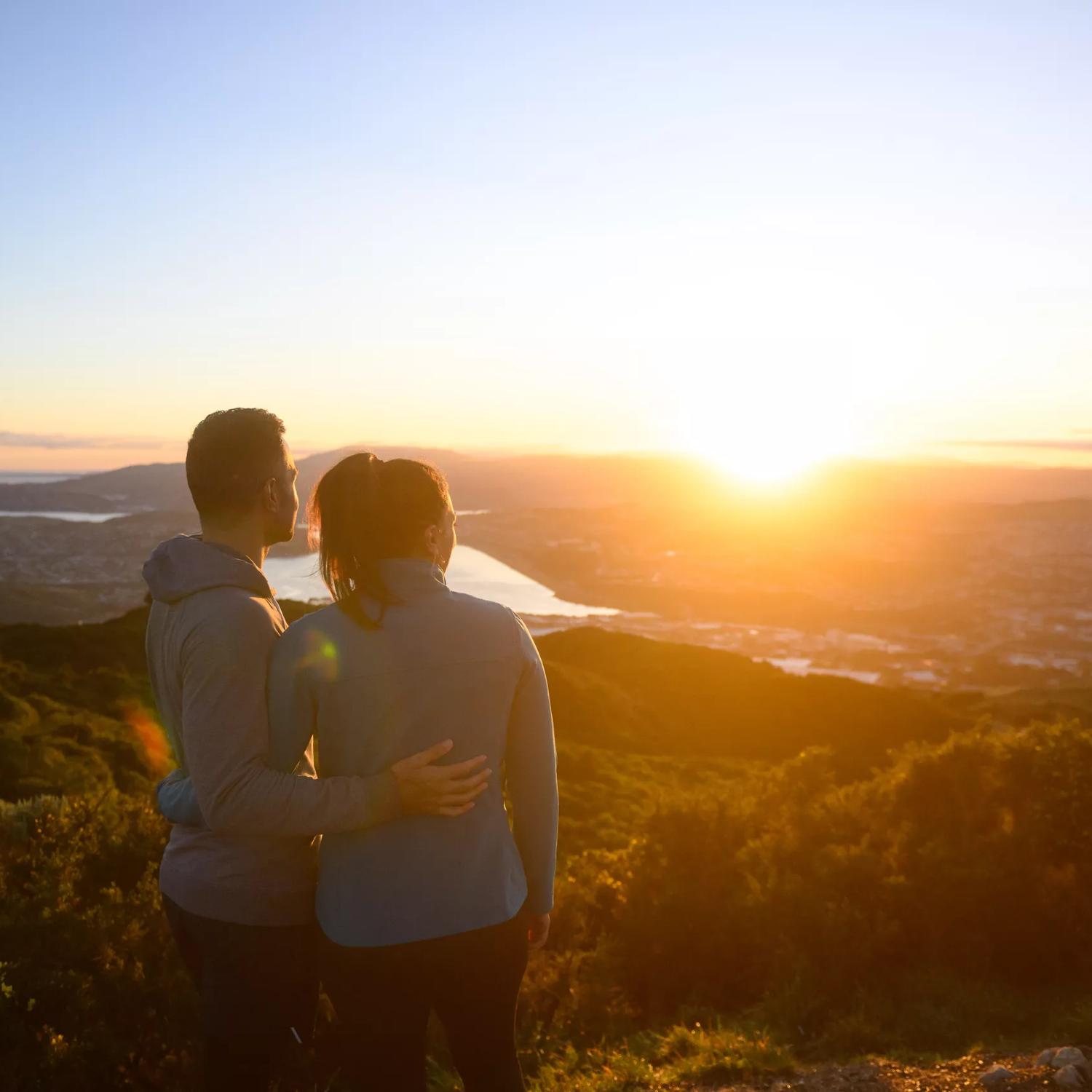 Two people stand arm in arm looking at a picturesque sunset from Mount Rangituhi, Colonial Knob Walkway. 