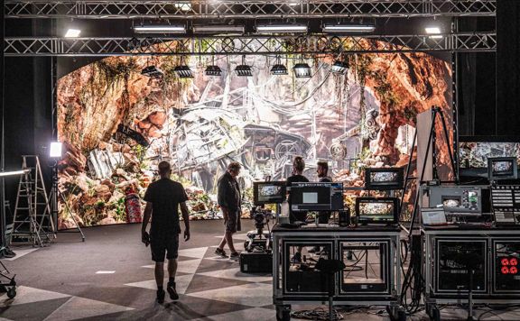 Behind the scenes on a film set with four crew members, a projection screen, lights and other film equipment.