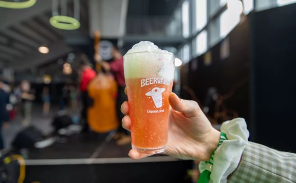 Close-up of a hand holding up a Beervana glass filled with an orange drink and foam atop.