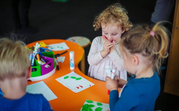 Young children at a bright orange table at Capital E, glueing paper leaves onto pieces of paper.