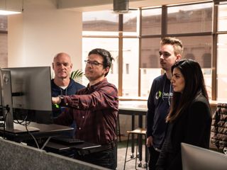 Four people in an office looking at a computer screen at a standing desk.