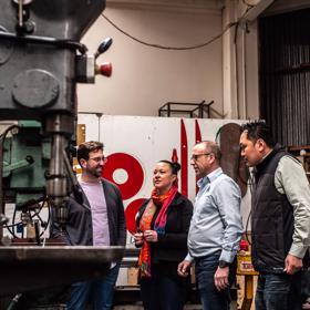 Four people stand inside a workshop at the Metal Art Ltd., a commercial furniture manufacturer located in Lower Hutt.