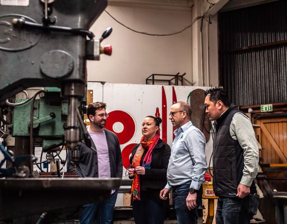 Four people stand inside a workshop at the Metal Art Ltd., a commercial furniture manufacturer located in Lower Hutt.