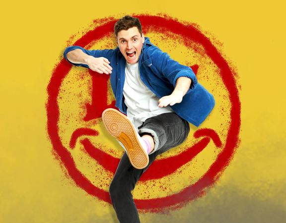 Luke Kidgell in a denim shirt, kicking one leg up towards the camera. Image is superimposed on a yellow background with a red spray painted smiley face.